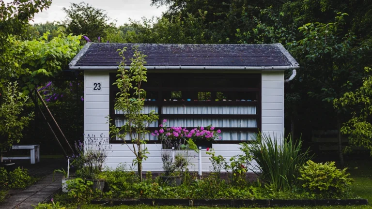 A small white wooden garden house with a dark roof surrounded by lush green foliage and flowers.