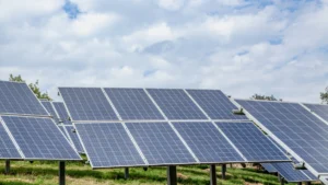 A large-scale array of solar panels installed in a field under a bright, cloudy sky.