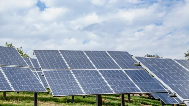 A large-scale array of solar panels installed in a field under a bright, cloudy sky.