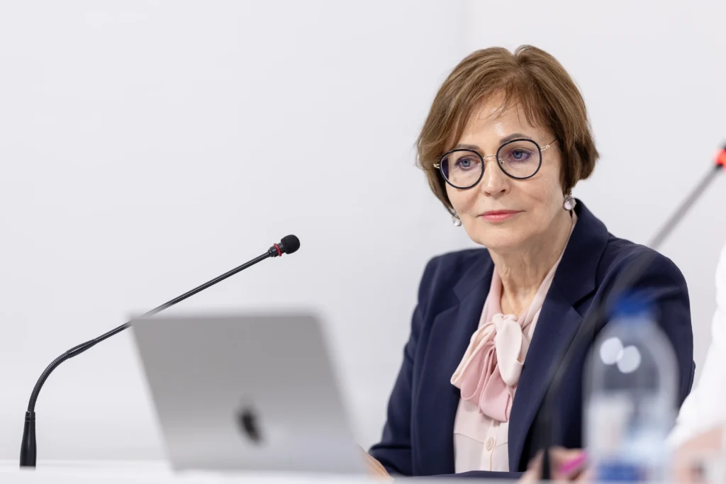 Woman wearing glasses and navy blazer sitting at a desk with a microphone and laptop.