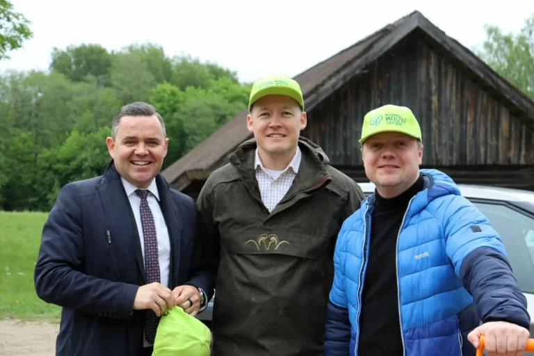 Three men smiling outdoors, two in lime green caps, standing before a rustic wooden barn.