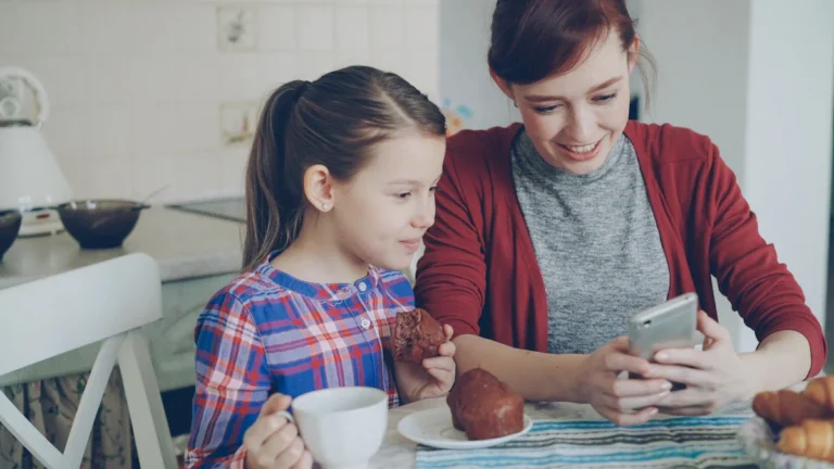 A mother and young daughter sit at a kitchen table looking at a smartphone together during breakfast.