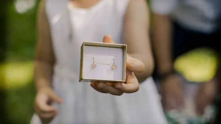 A young girl in a white dress holds out a small box containing heart-shaped earrings.