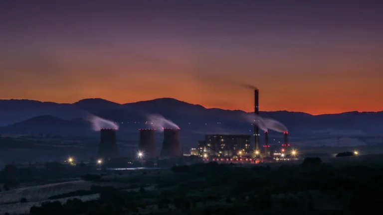 A cargo ship sails on water at sunset with the silhouette of a power plant background.