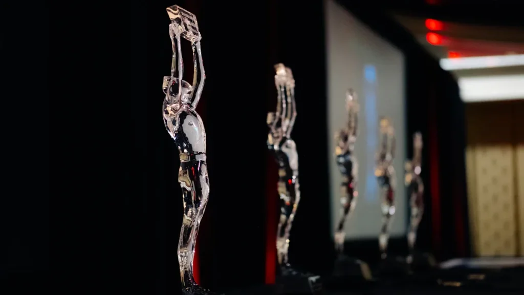 A row of clear glass award statuettes standing on a black table against a dark background.