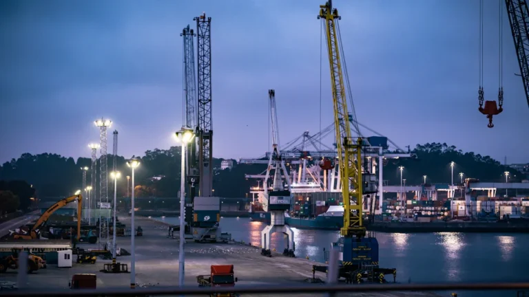 A modern industrial shipping port at dusk with illuminated cranes and infrastructure reflecting in the water.