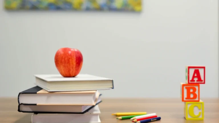 A red apple rests on a stack of books alongside colored pencils on a wooden desk.