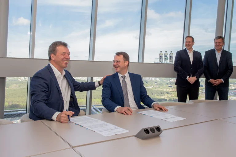 Two men in suits laughing while signing documents at a conference table with a city view.