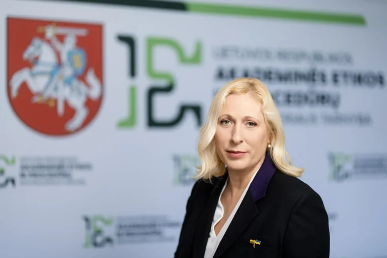 Blonde woman in professional attire standing before a Lithuanian government background and coat of arms.