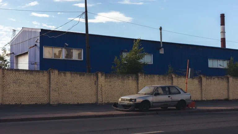 A silver car with a damaged, hanging front bumper parked on a roadside in an industrial area.