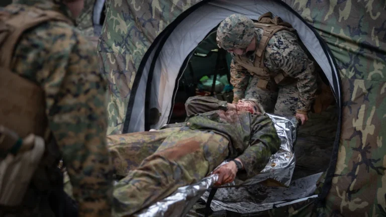 Military medics transporting a simulated casualty on a stretcher through a field environment.