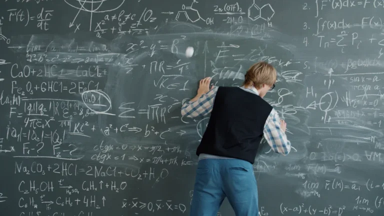 A man writing complex mathematical and scientific formulas on a large, crowded chalkboard in a classroom.