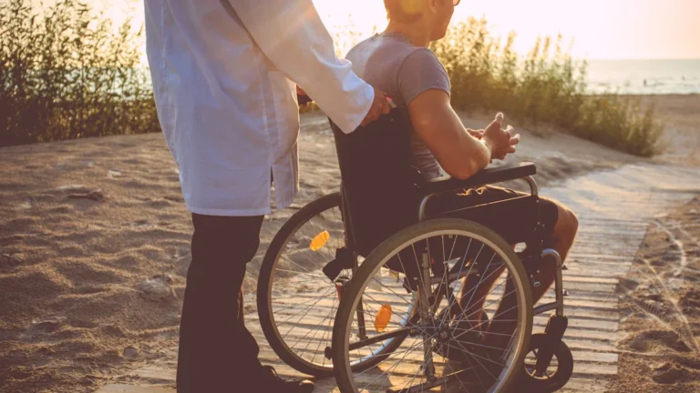 A professional in a white coat assisting a man in a wheelchair on an accessible wooden path at a beach.
