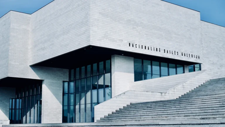 A modern white stone building with large glass windows and Lithuanian text under a blue sky.