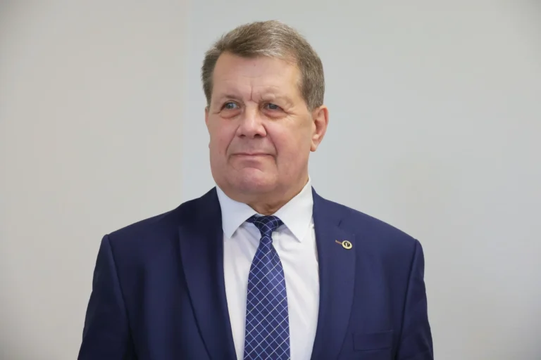 A man wearing a dark blue suit and patterned tie against a plain white background.