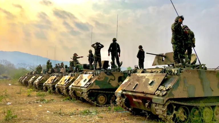 A line of armored personnel carriers with soldiers standing on top in a rural field setting.