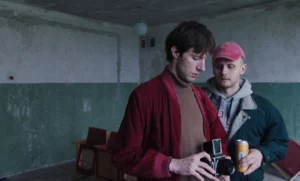 Two men in a derelict room, one holding a vintage twin-lens camera while another watches.