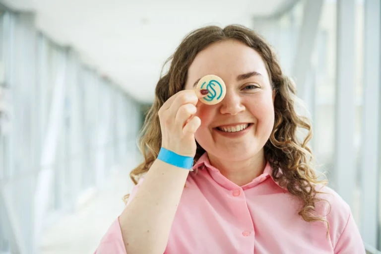 Smiling woman with curly hair holding a round cookie with a blue logo over her eye.