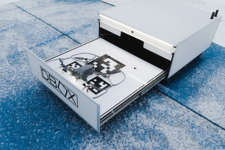 A small drone sits on a patterned landing pad inside an open white automated docking station.