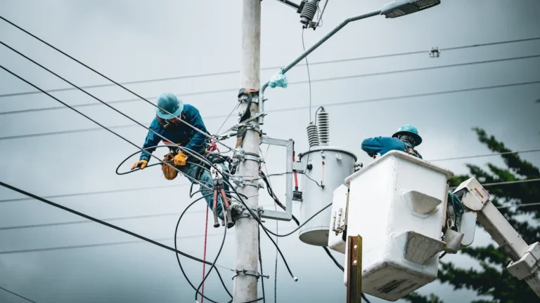 Utility workers in hard hats and safety gear working on a power pole with complex wiring and a transformer.