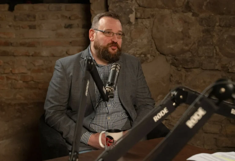 Bearded man wearing glasses and a blazer speaking into a podcast microphone in a studio.