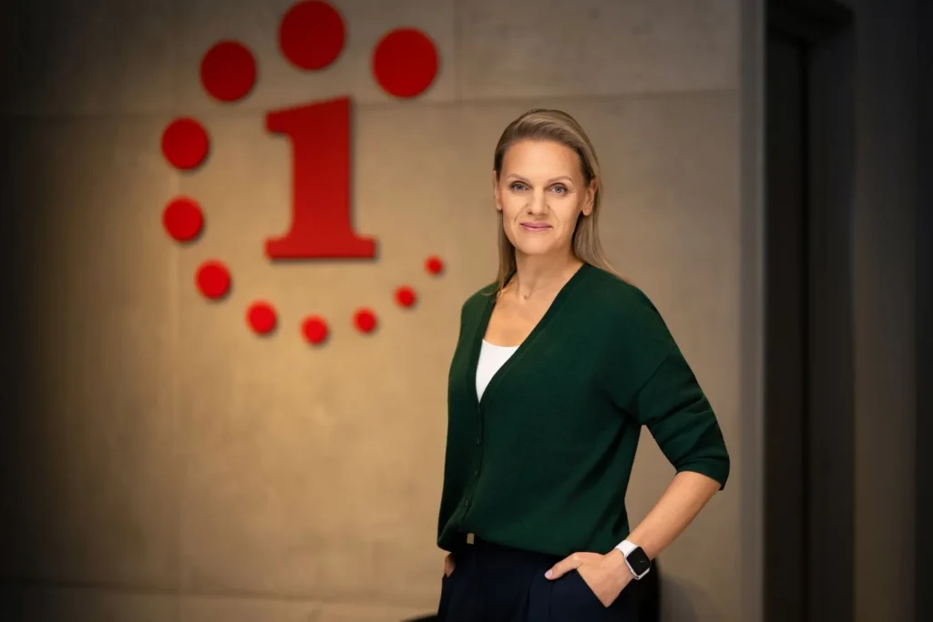 A woman wearing a green cardigan poses in front of a red number one logo.