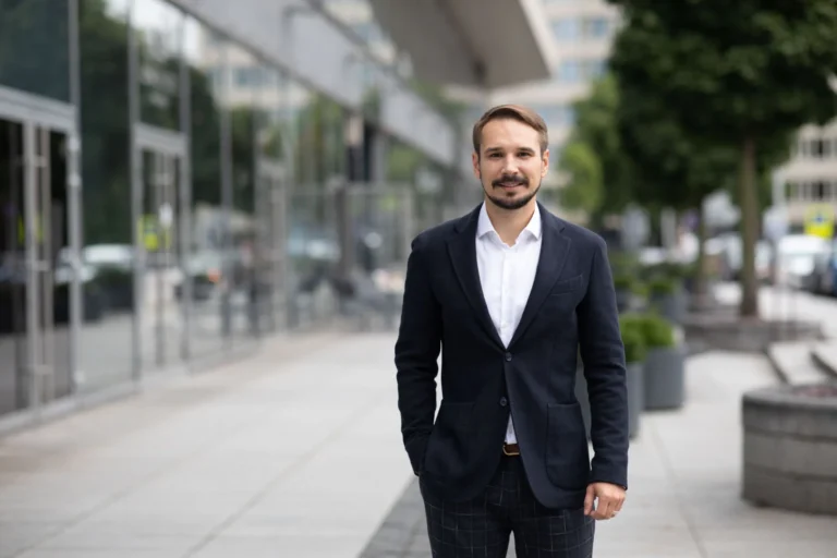 Man with beard wearing a navy blazer and white shirt standing on a city sidewalk.