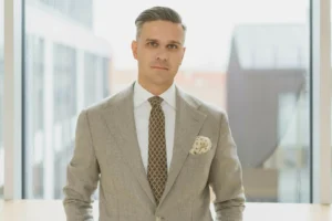 A man wearing a grey suit, white shirt, and patterned tie stands before a bright window.