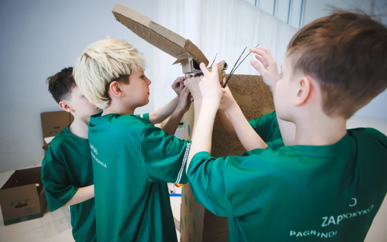 Three young boys in matching green t-shirts work together to assemble a large cardboard sculpture.