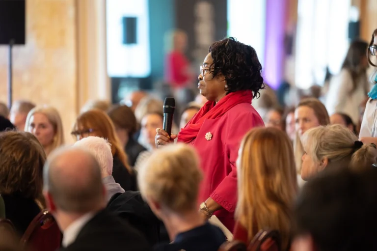 A Black woman in a pink blazer and scarf holding a microphone at a public conference.