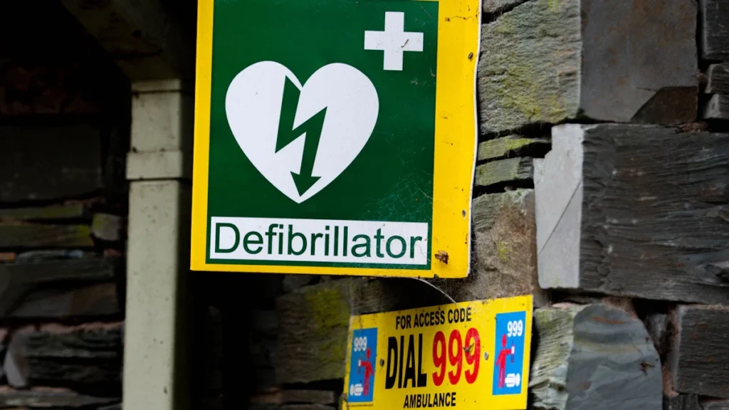 A green and yellow emergency sign for a defibrillator mounted on a stone wall in the UK.