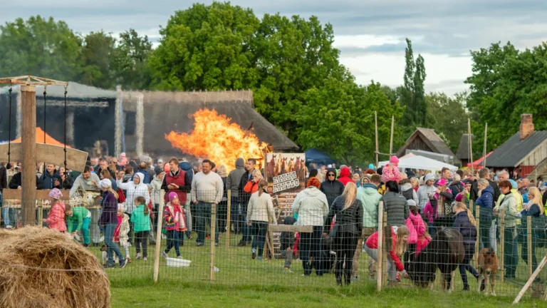 A festive crowd gathers around a large traditional bonfire near rustic wooden buildings during a solstice celebration.