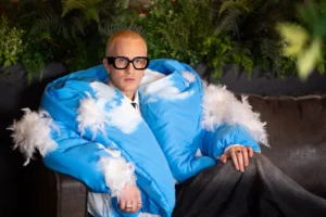 Man in glasses wearing a voluminous blue cloud-print jacket with white feathers on a couch.