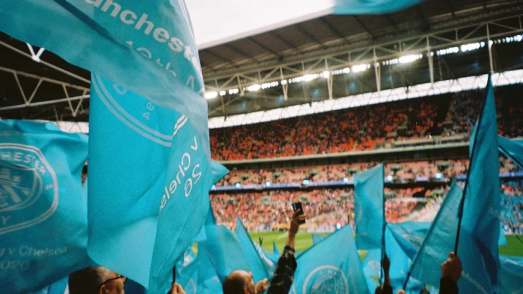 Fans waving light blue flags in a crowded stadium with a large arched roof structure overhead.