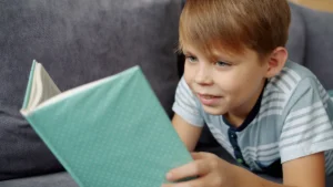 A young boy smiling while reading a book on a grey sofa at home.