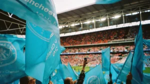Supporters wave sky blue Manchester City flags in a crowded stadium during a match.