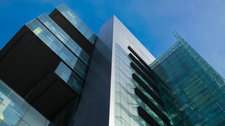 A low-angle shot of a modern glass and steel building against a bright blue sky, representing the urban landscape of Manchester.