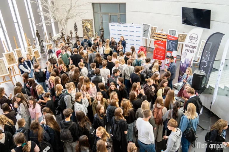 Crowded indoor hall with young people standing near corporate banners and artistic displays on easels.