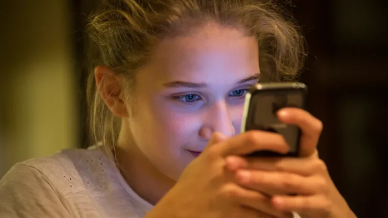 A young girl's face illuminated by the blue glow of a smartphone screen in a dimly lit environment.