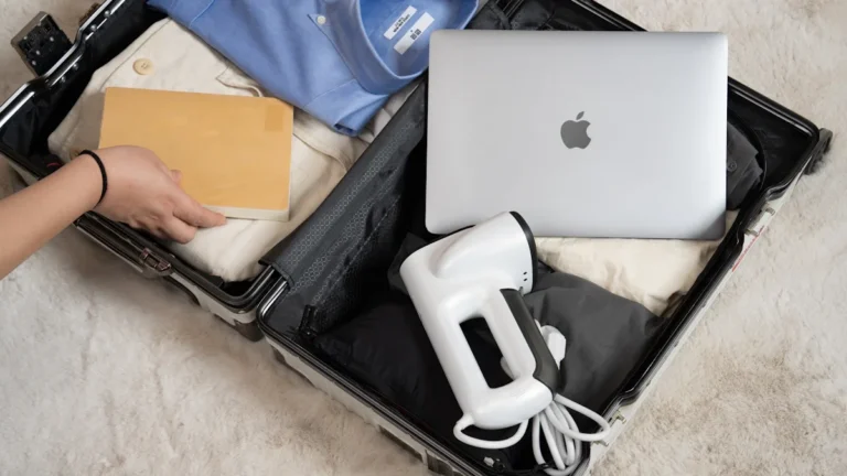 An open suitcase on a neutral rug containing a blue dress shirt, a laptop, and travel essentials.