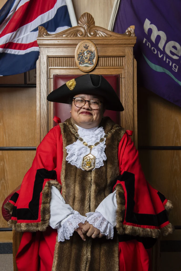 Person wearing traditional red mayoral robes and tricorn hat sitting in an ornate wooden chair.