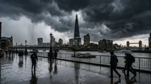Pedestrians with umbrellas walk along a wet London riverfront under dark, stormy clouds.