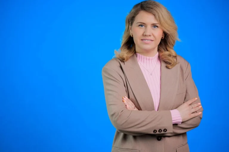A blonde woman in a tan blazer and pink top posing against a blue background.