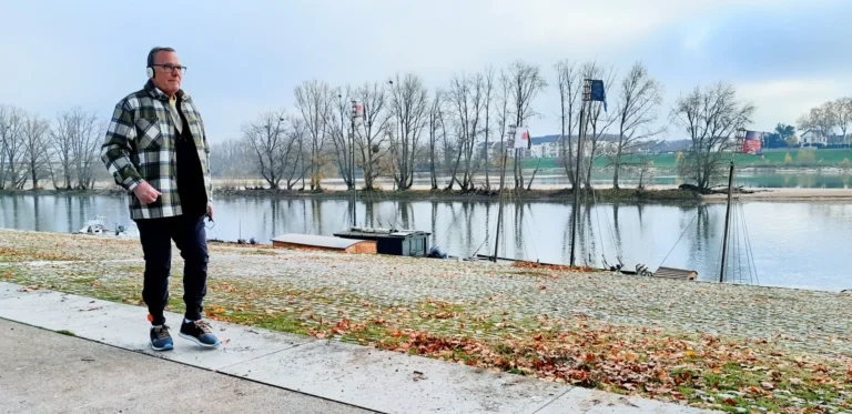Senior man in a plaid jacket and headphones walks along a riverbank with moored boats.
