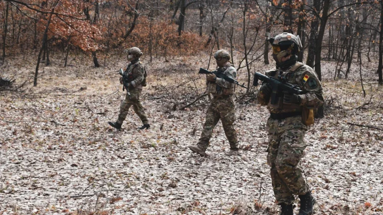 Three soldiers in tactical camouflage gear patrol through a wooded area during a NATO military exercise.