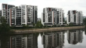 Contemporary multi-story apartment buildings with glass balconies reflecting in a calm river under an overcast sky.