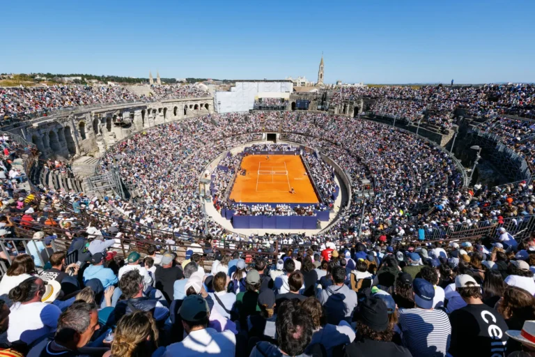Aerial view of an orange clay tennis court inside a packed, sunlit ancient stone amphitheater.