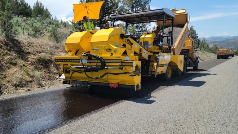 A specialized yellow road surfacing vehicle applies a fresh coat of asphalt to a rural road.