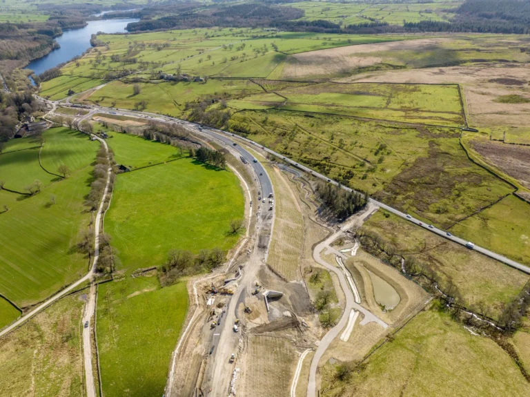 Aerial view of heavy machinery and road construction progressing through green rural fields and hills.