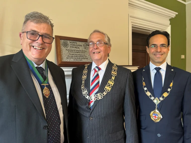 Three men in suits and ceremonial chains stand before a North Yorkshire Council commemorative plaque.